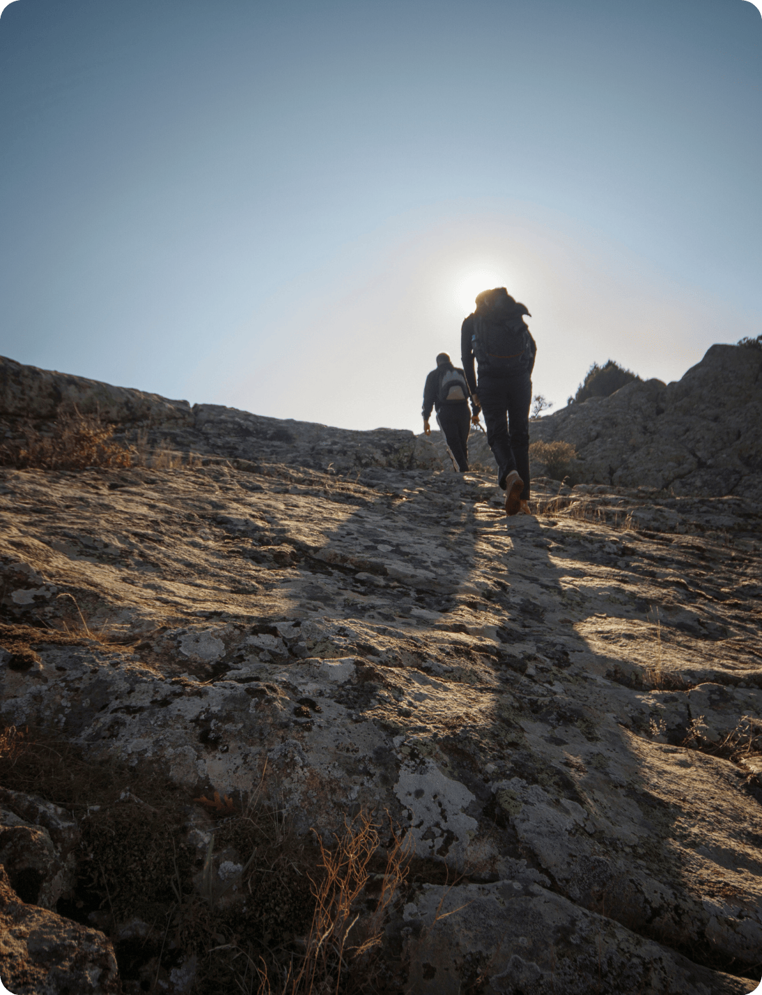 Two hikers with backpacks climb a rocky slope, casting long shadows in the sunlight, with the sun positioned just above their heads in a clear sky.