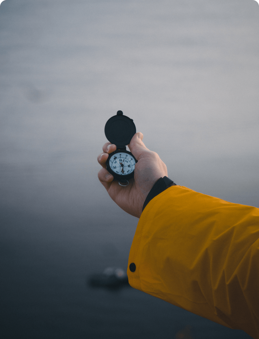 A person wearing a yellow jacket holds an open compass in their hand, pointing it toward a calm, gray body of water in the background.