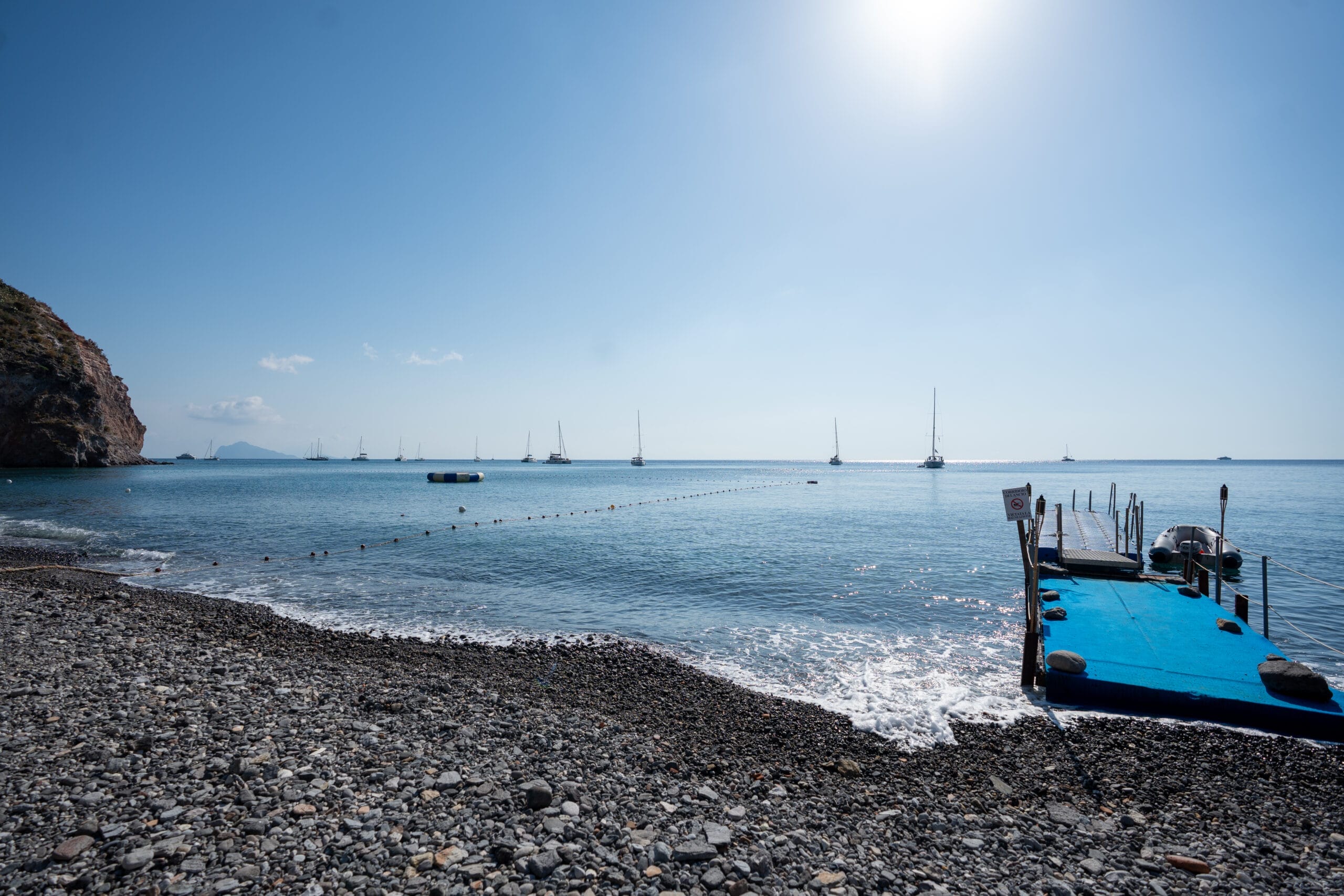 A calm, pebbly beach with a floating blue dock extends into the sea, perfect for community event sailing. Several sailboats are anchored under a clear blue sky and bright sun, with a rocky cliff visible on the left.