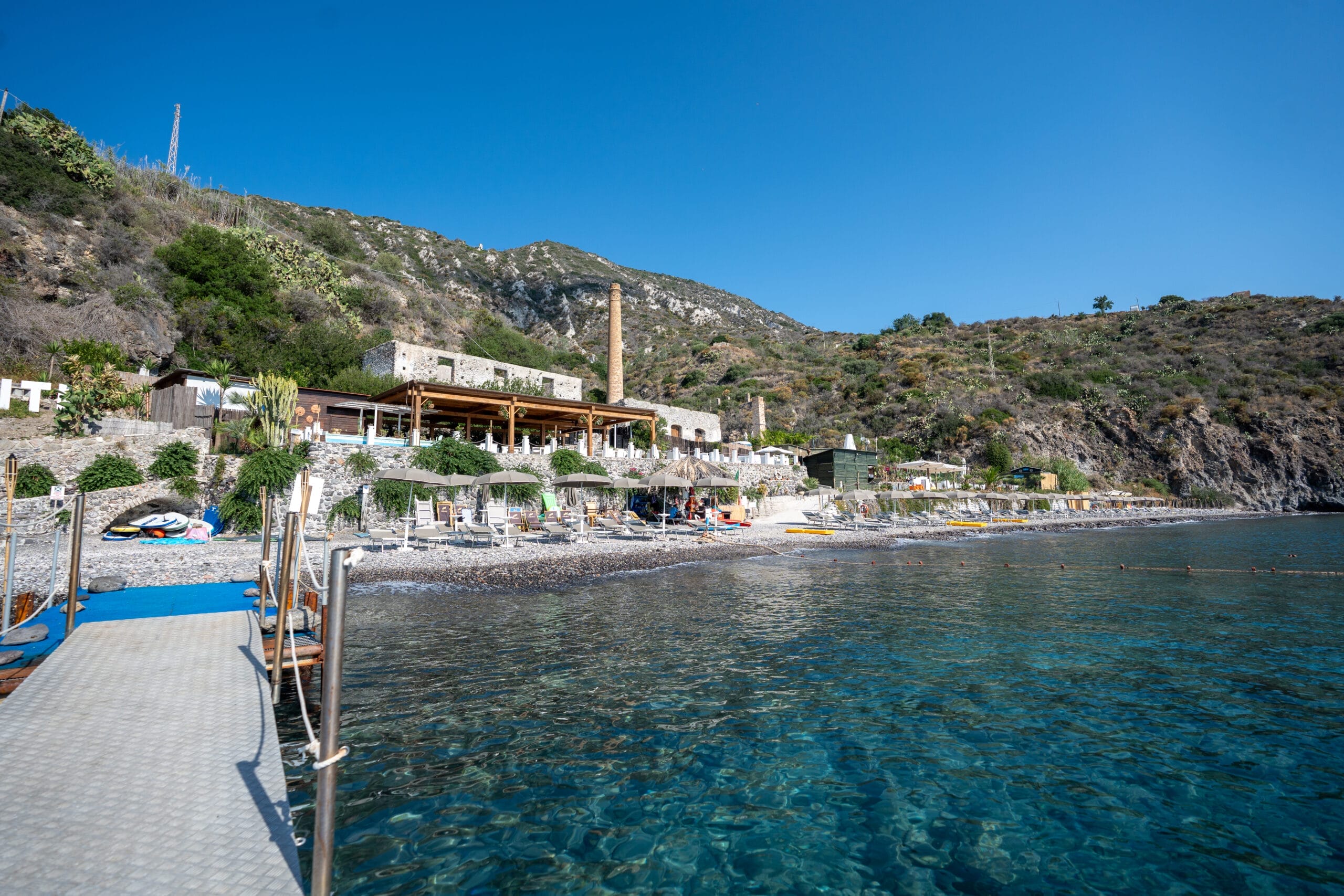 A rocky shoreline with clear blue water, a dock leading to a beach area with lounge chairs and umbrellas, and stone buildings nestled against a hillside under a clear blue sky—perfect for the unique Sailing Turkey 2026 experience.