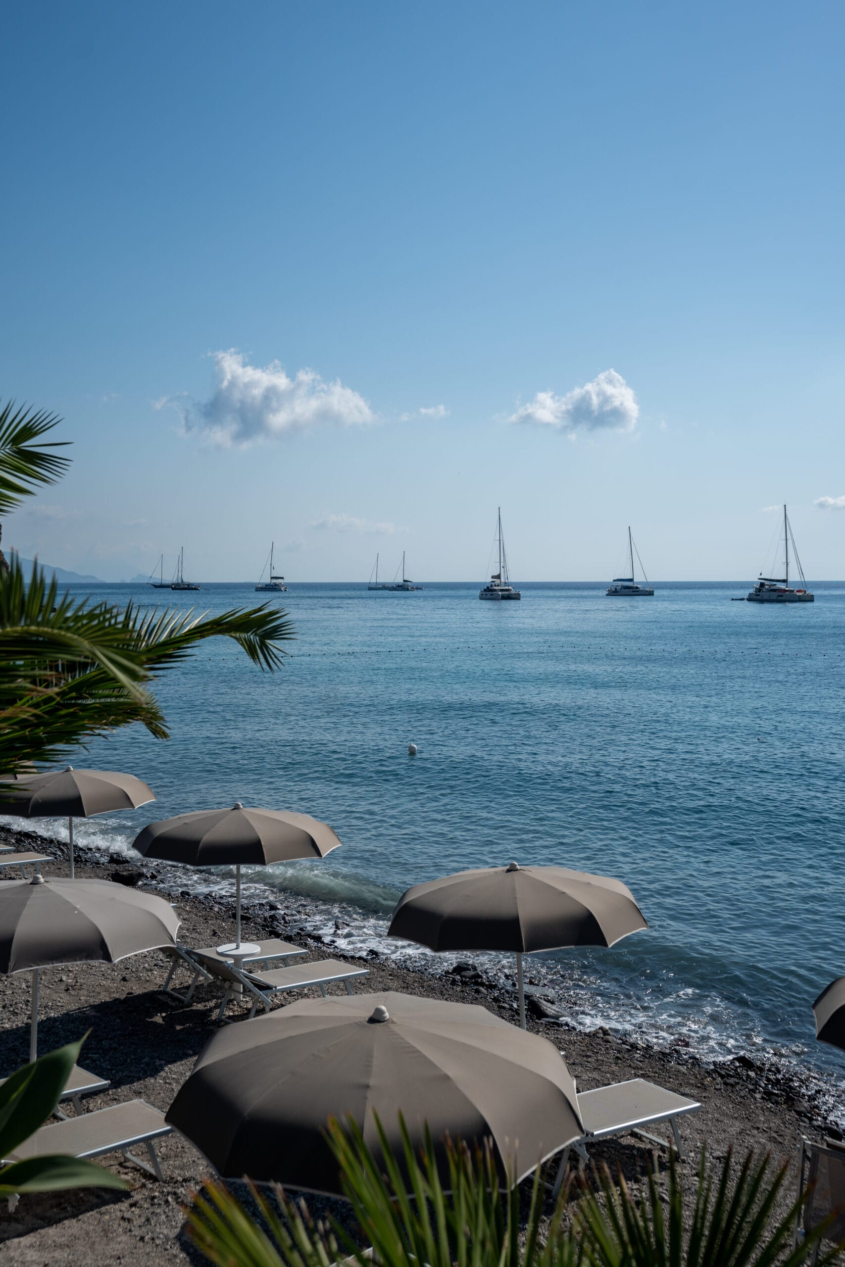 A tranquil beach scene with gray umbrellas and lounge chairs on pebbles, overlooking sailboats floating on calm blue water under a clear sky—perfect for dreaming of Sailing Turkey 2026. Palm leaves gracefully frame the inviting view.