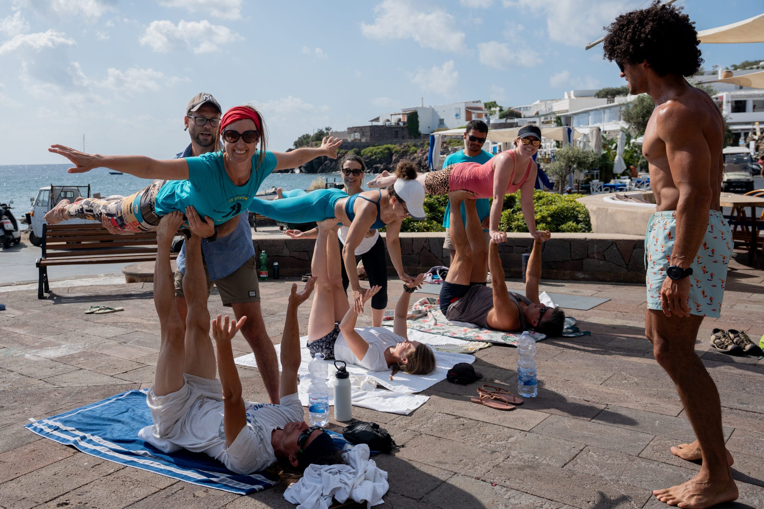 A group of people practice acroyoga outdoors near the coast during a lively community event sailing, with some balancing others in the air while others watch or assist. The sunny scene features buildings and the sea in the background.