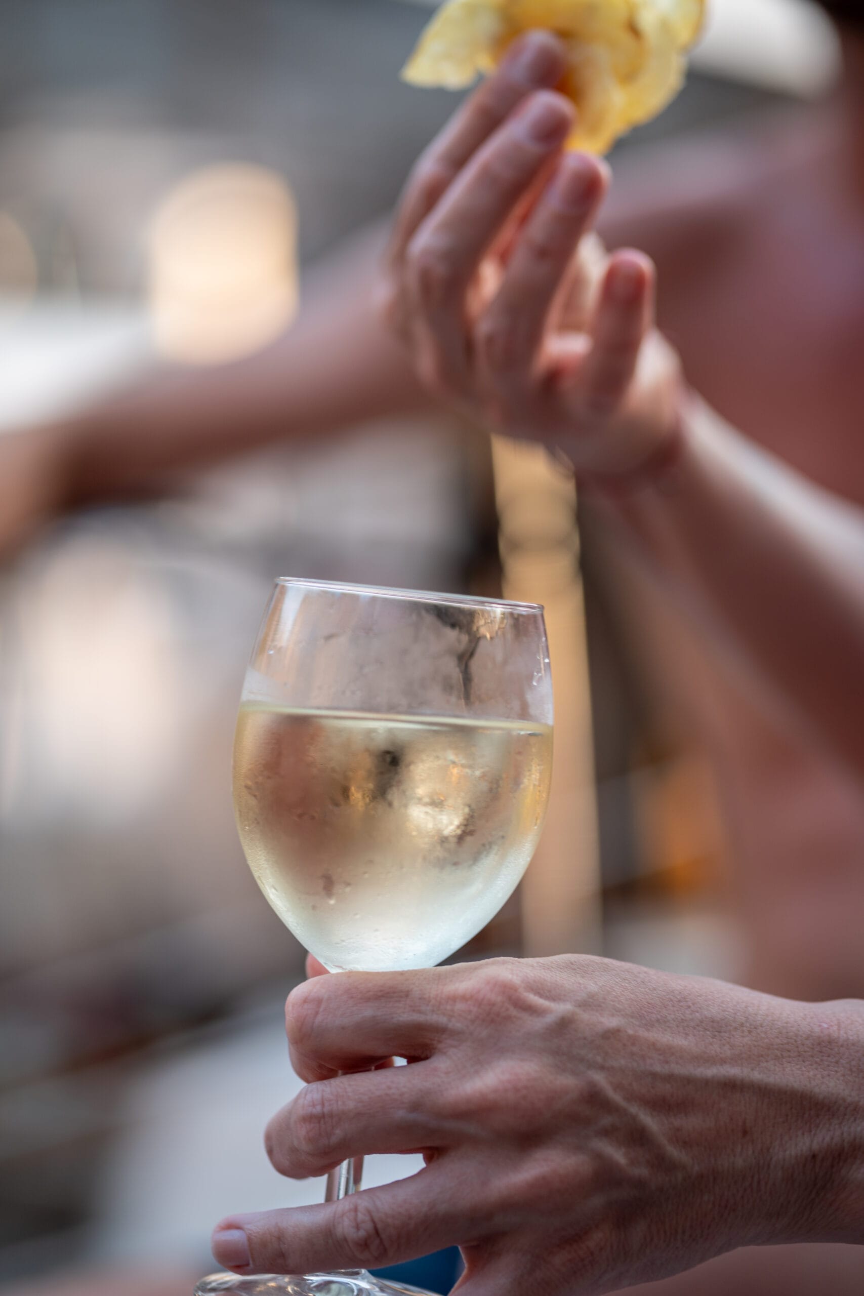 A close-up of a person holding a glass of chilled white wine, with another hand in the background holding a crisp potato chip—capturing the relaxed vibe of a community event sailing on Turkey’s coast during Adventure Sailing Week 2026.