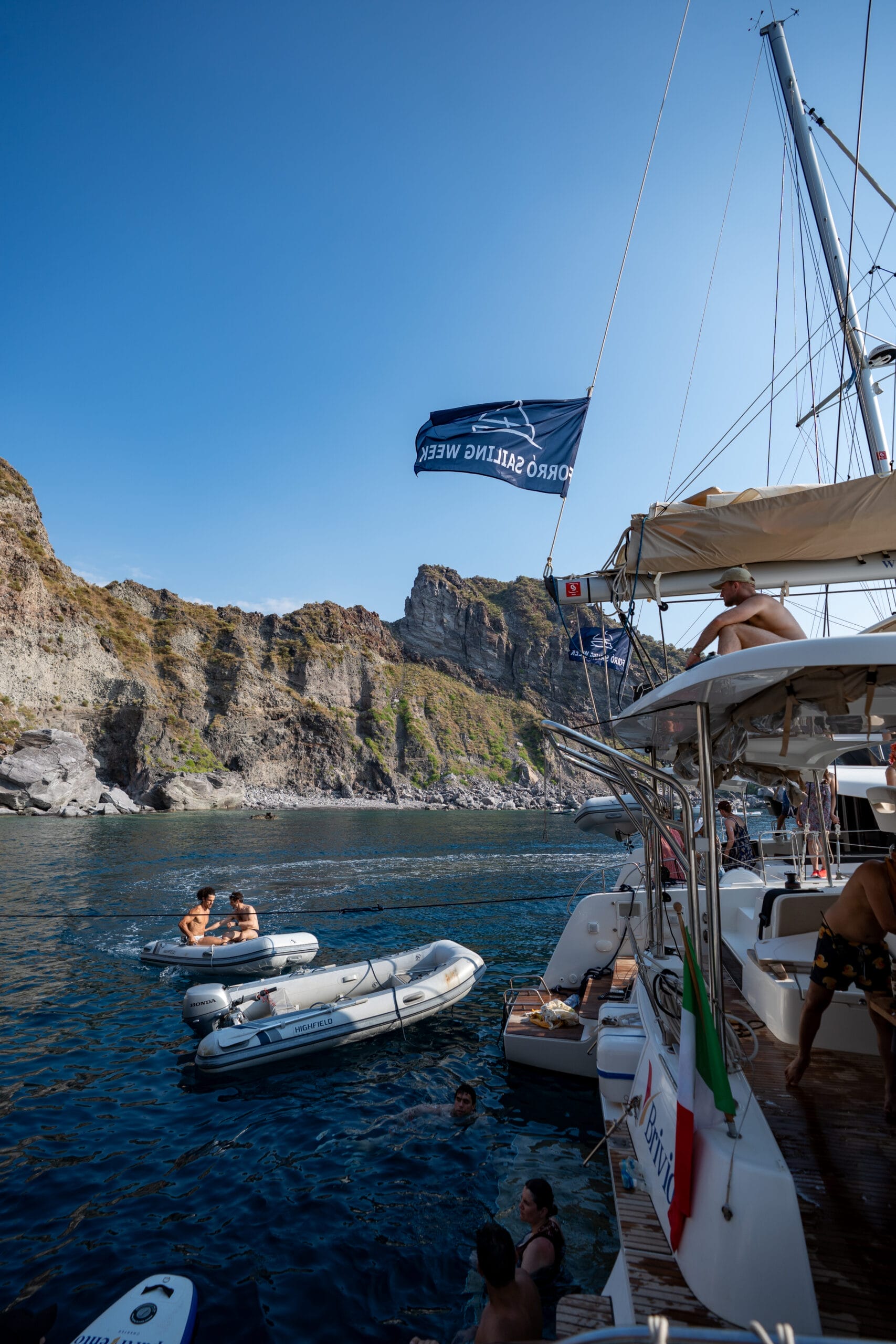People are relaxing on a yacht and in small inflatable boats on calm blue water near rocky cliffs during a community event sailing. A flag reading "SORAO SAILING WEEK" flies, showcasing the unique sailing Turkey experience under sunny skies.